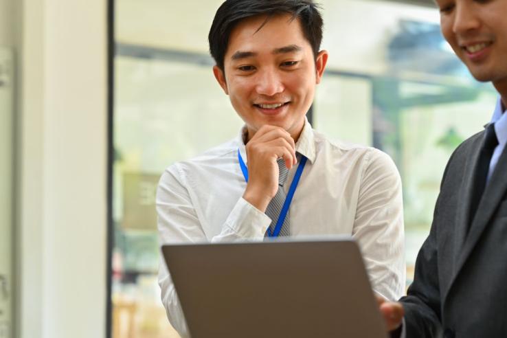 Two Asian men in formal business clothing are engaged in a discussion while viewing a laptop together in a workplace.